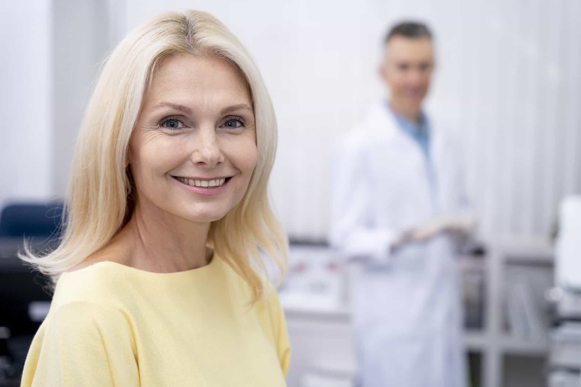 middle aged blonde pretty woman smiling with dentist in lab coat in the background